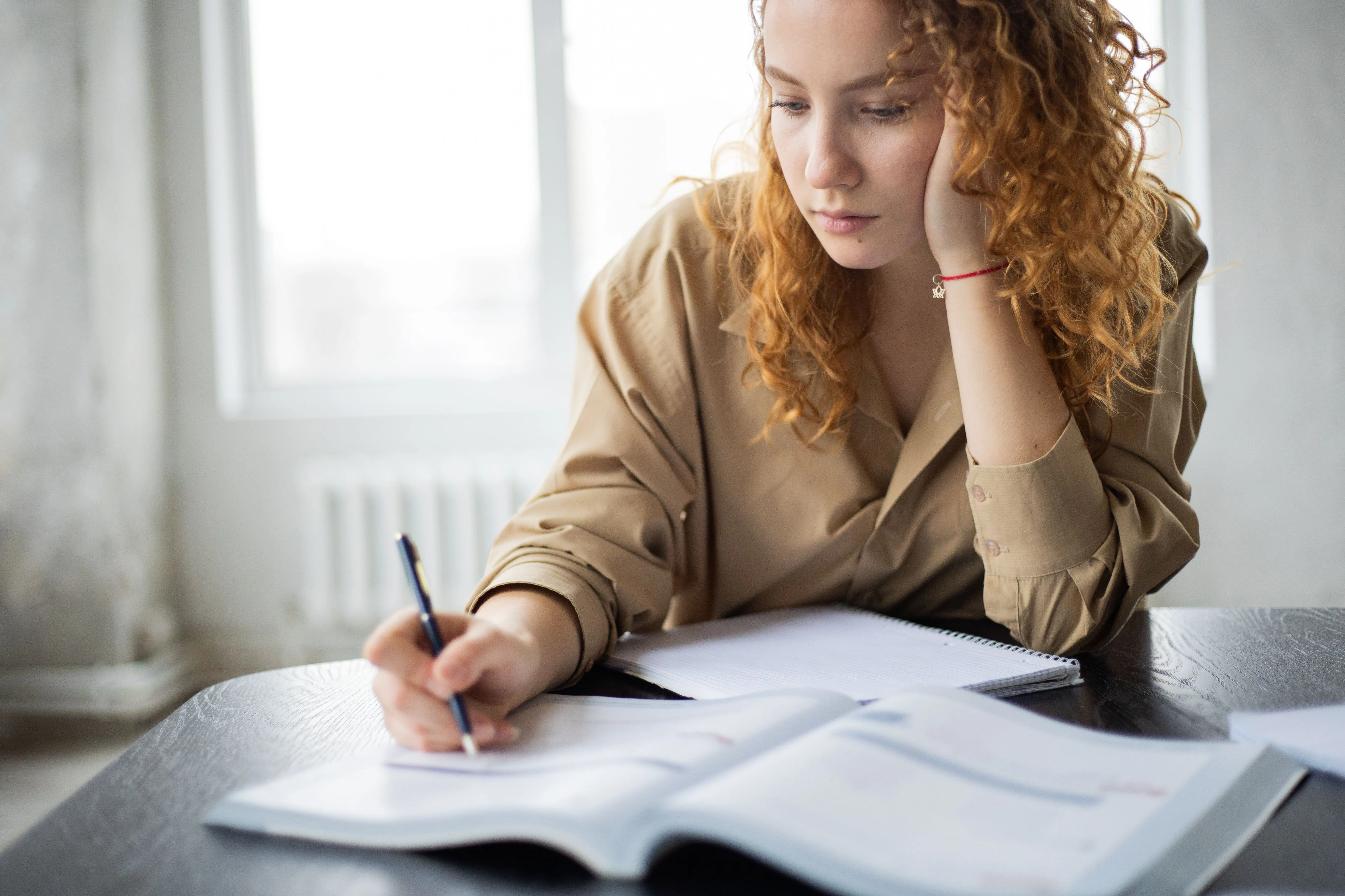 Mulher inclinada sobre caderno com anotações, cotovelo apoiado na mesa, luz natural ao fundo